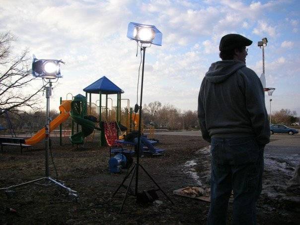 Two tall and bright hot lamps are standing on a playground. The play place is in the background. One man stands in the foreground, facing away from camera